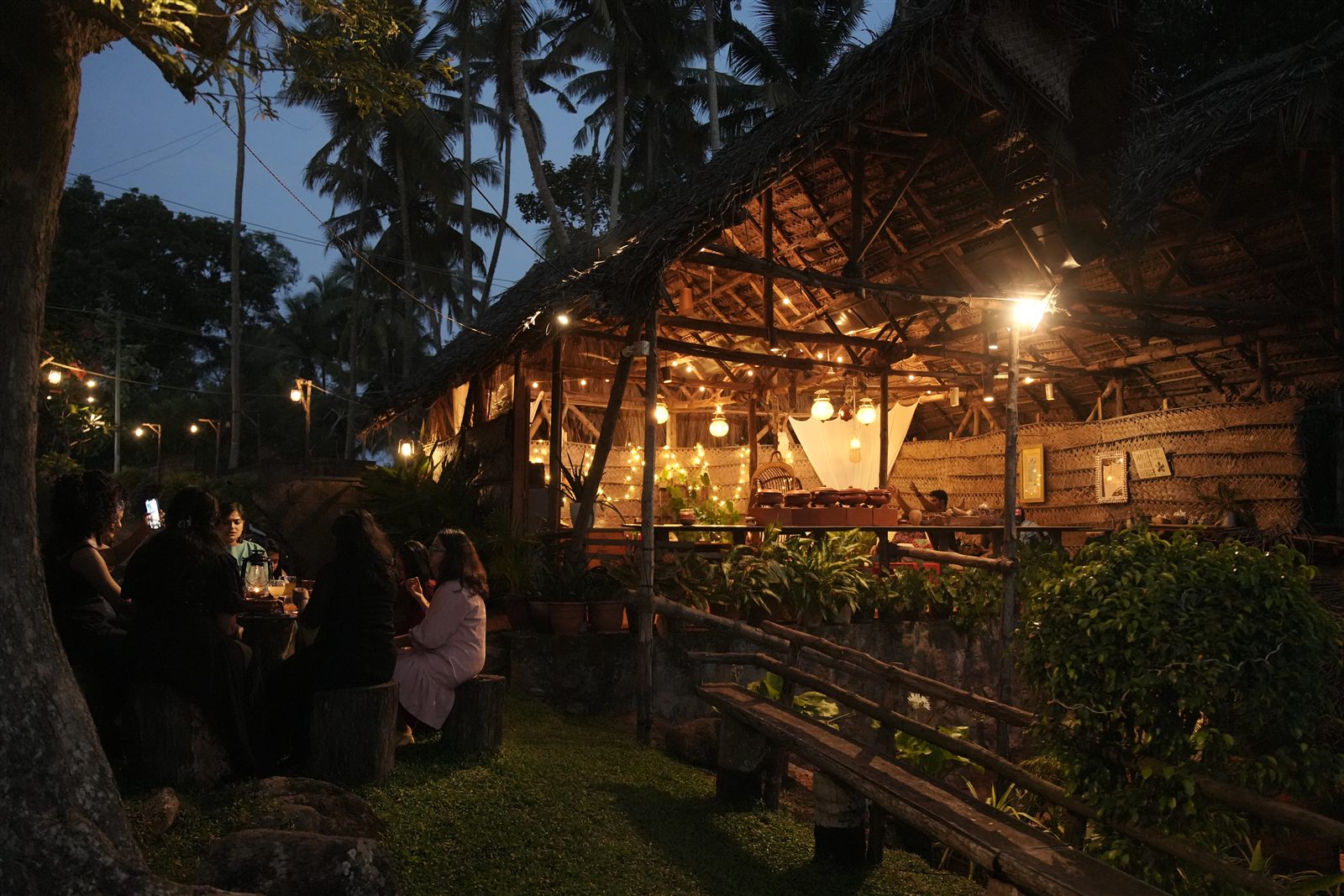 The palm thatch pavilion glowing at dusk, guests at a lantern lit table under the big tree